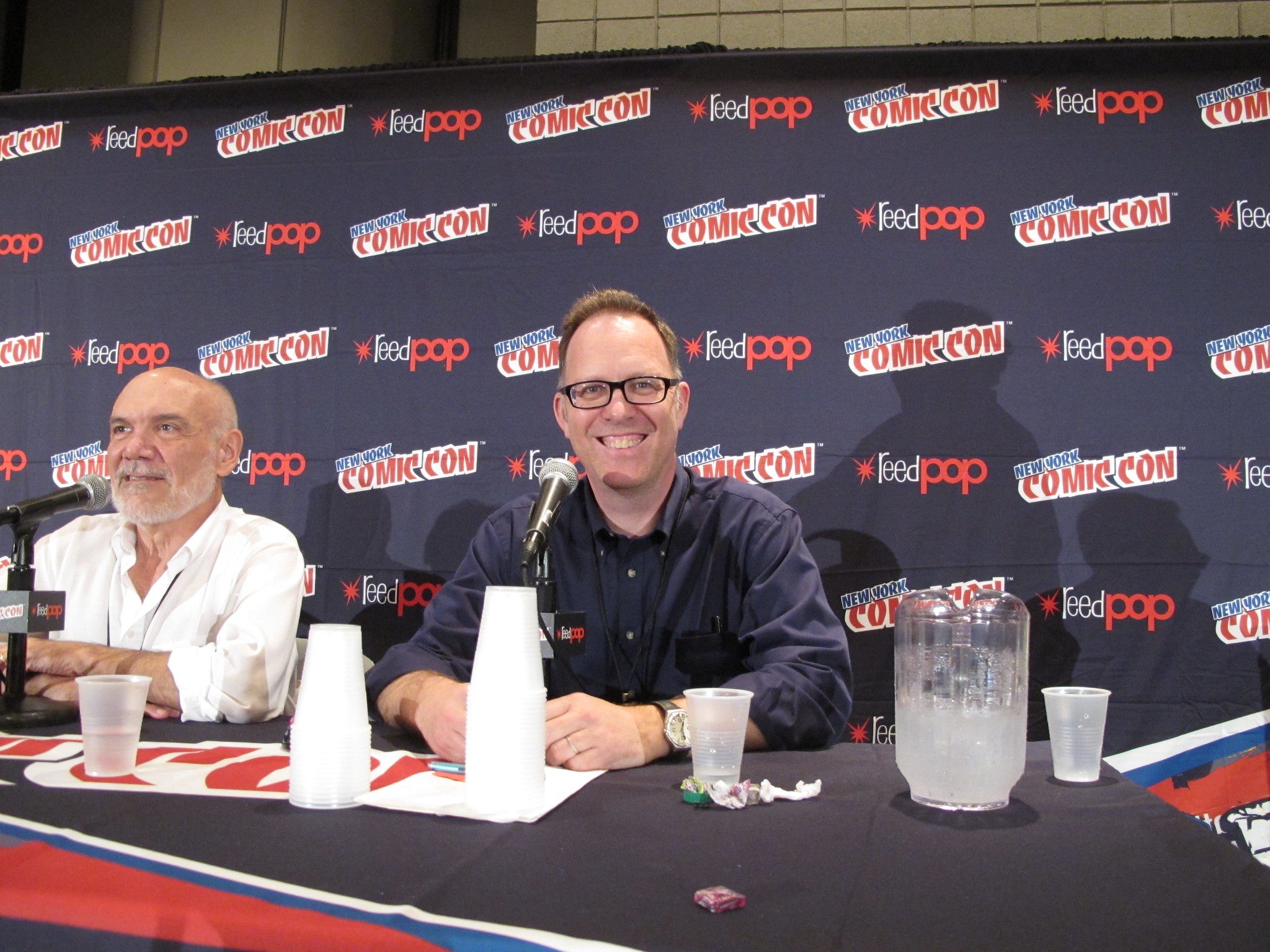 Two men sitting at a table with a Comic Con backdrop