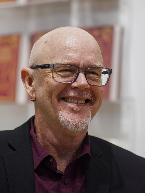 A smiling bald man with glasses, a goatee, and a small hoop earring, wearing a dark blazer over a dark purple collared shirt, photographed indoors against a softly blurred background.