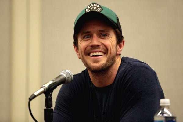 Man wearing a green cap with text, sitting in front of a microphone at an event.