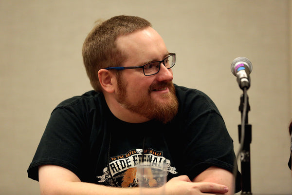 Man with glasses and a beard sitting in front of a microphone at an event.