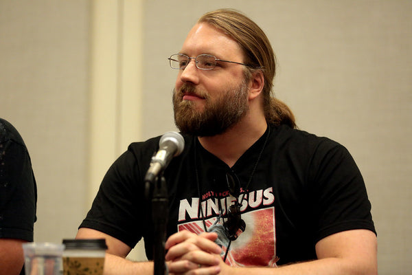 Man with long hair and beard wearing a black t-shirt with a graphic design, sitting in front of a microphone.