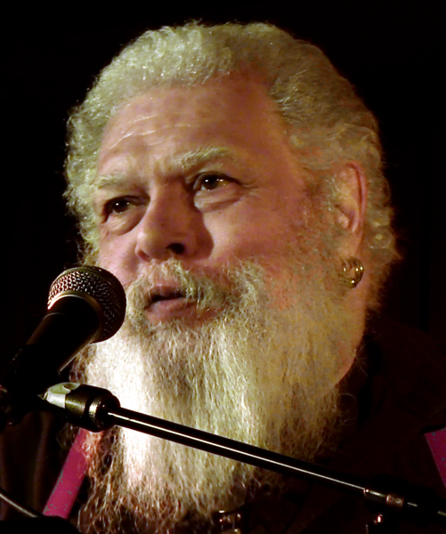 Man with a beard speaking into a microphone against a dark background