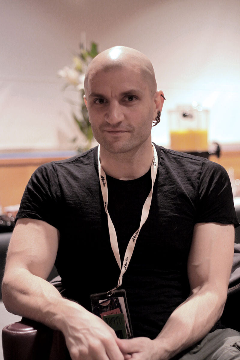 Man with a shaved head wearing a black t-shirt and lanyard, sitting indoors.