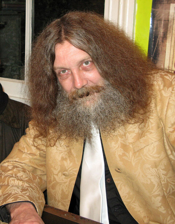 Man with long hair and beard wearing a patterned beige jacket indoors.