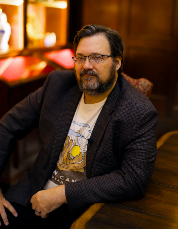 Man wearing glasses and a dark jacket over a graphic t-shirt in a dimly lit room.