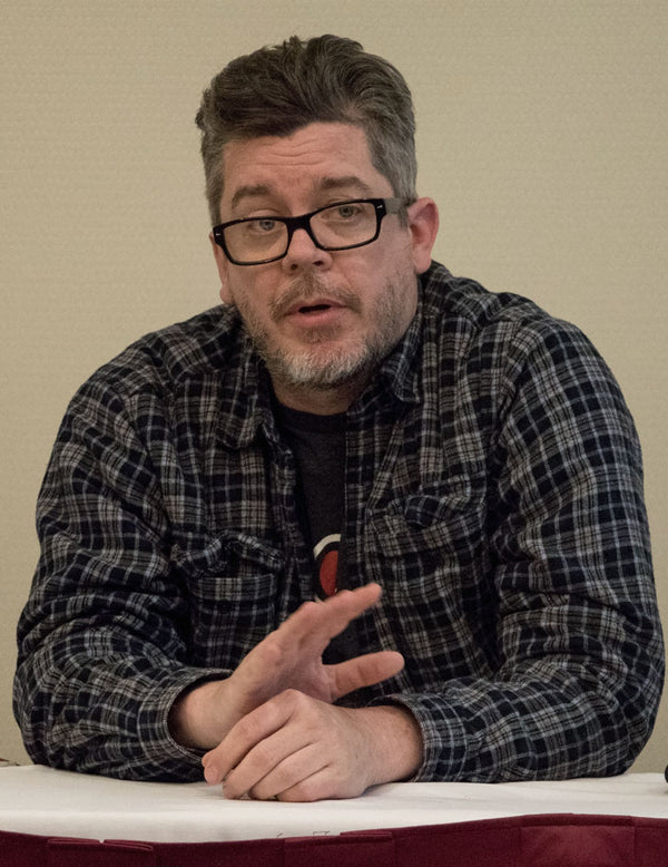 Man wearing glasses and a plaid shirt sitting at a table with a beige background