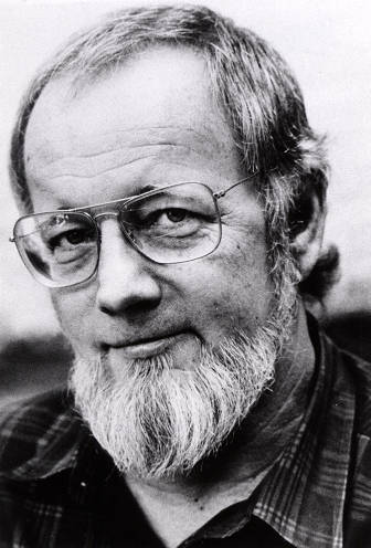 A black and white close-up portrait of an older man with a white beard, wearing wire-rimmed glasses and a plaid shirt.