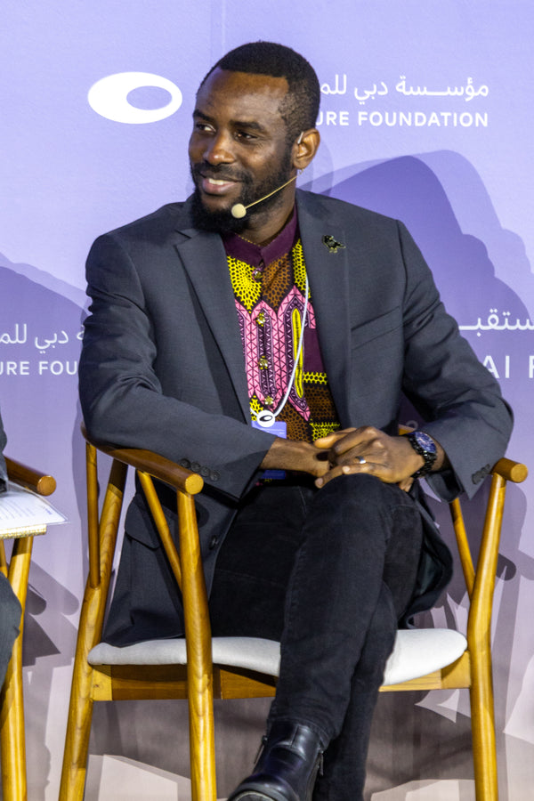 Man sitting on a chair at an event with a microphone, wearing a suit and colorful shirt.