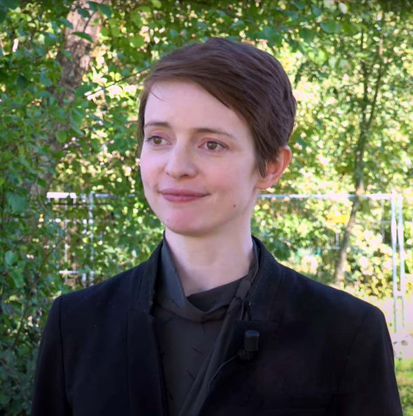 Person wearing a black coat standing outdoors with greenery in the background