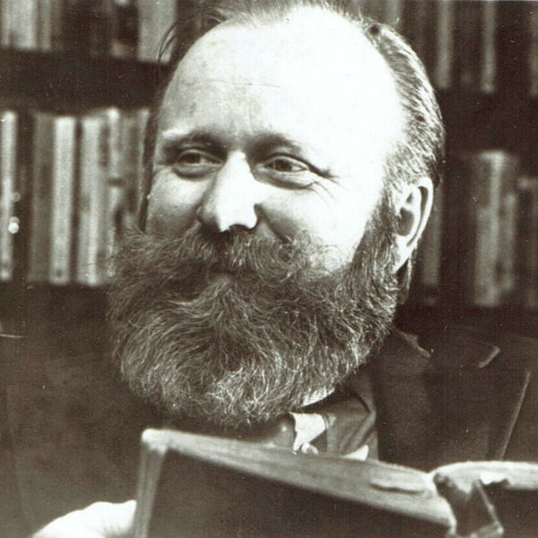 Black and white photo of a man with a beard holding a book in front of a bookshelf.