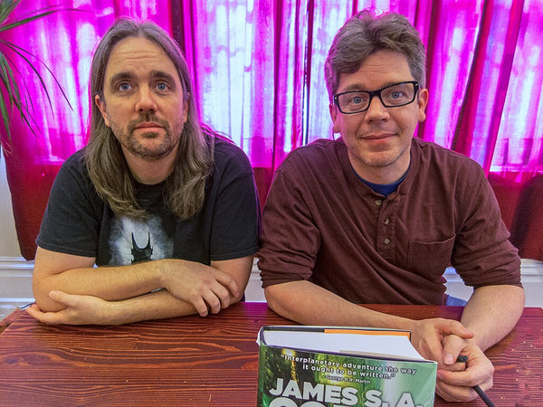 Two men sitting at a table with a book titled 'James S.A.' in front of pink curtains.