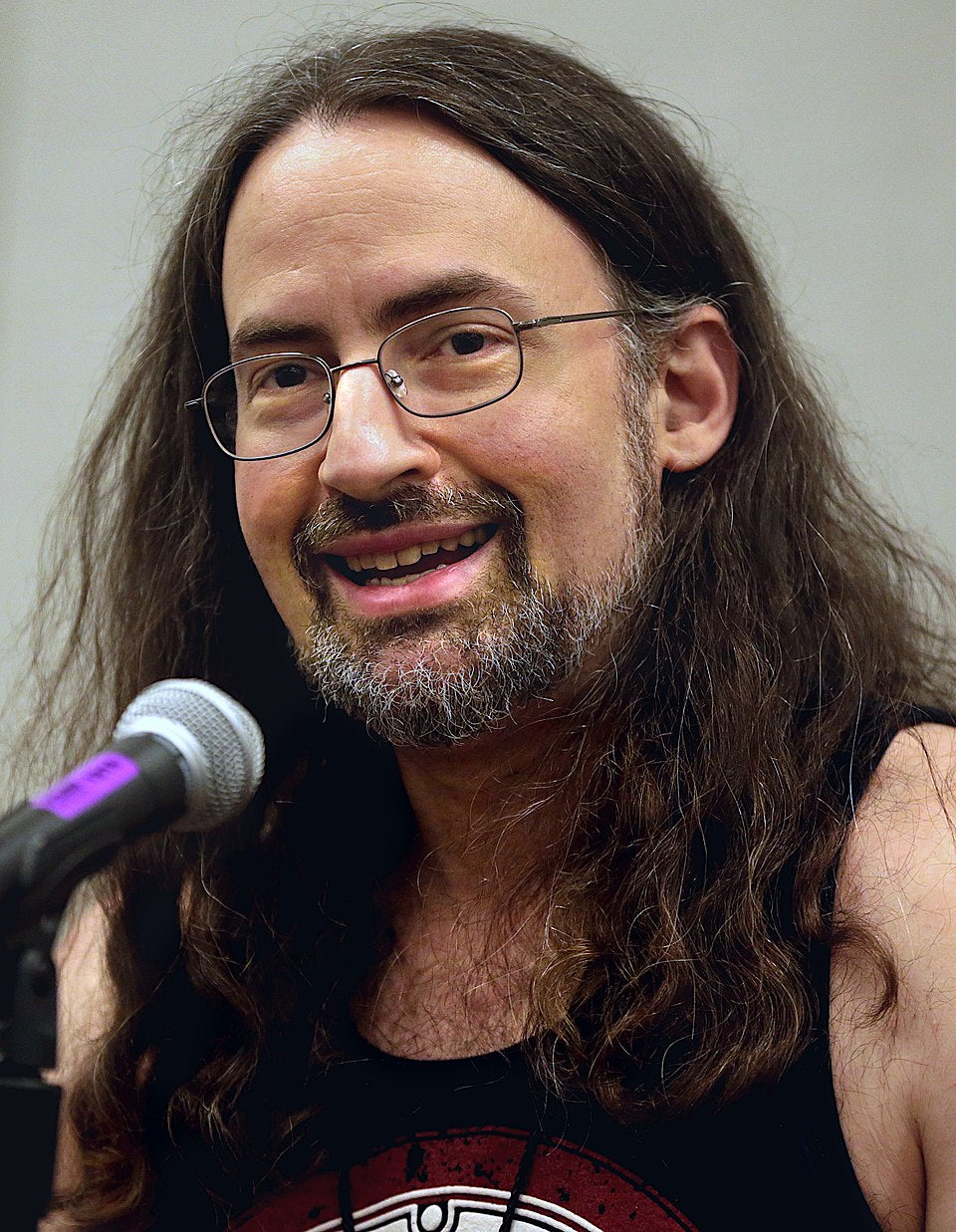 Man with long hair and beard speaking into a microphone against a plain background