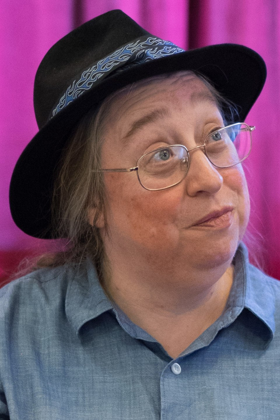 Person wearing a black hat with a blue band and glasses, against a pink curtain background