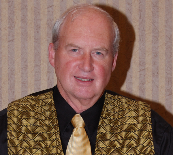 Man wearing a patterned vest and yellow tie against a striped wall.