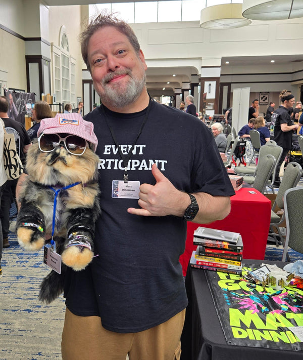 Man holding a small dog plushie with sunglasses, wearing an 'Event Participant' shirt in a convention center setting.
