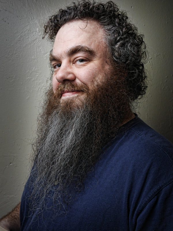 Man with a long beard and curly hair wearing a blue shirt against a plain background