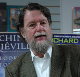 Man with a beard wearing a suit in an indoor setting with books and posters on the wall.