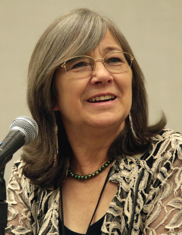 Woman with glasses speaking into a microphone against a plain background