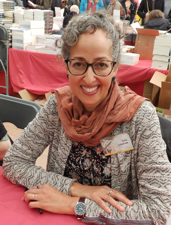 Woman with glasses and a scarf sitting at a table with books and people in the background