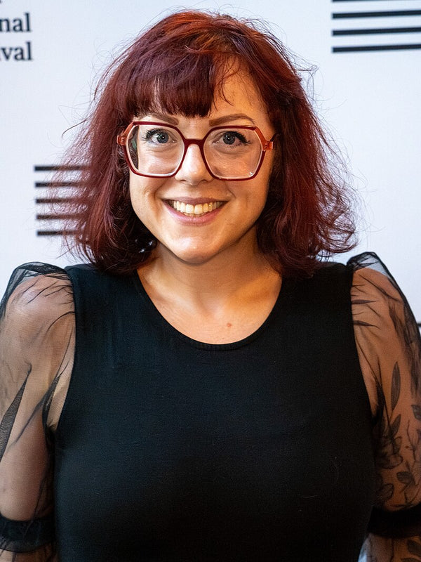 Person with red hair and glasses smiling in front of a blurred background