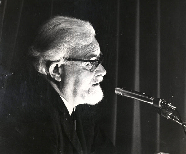 Black and white photo of a person with glasses speaking into a microphone.